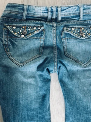 Coola utsvängda bootcut jeans med pärldetaljer - Snygga ljusblå jeans med slitningar och pärldetaljer på bakfickorna. Jeansen har klassisk femficksmodell, låg midja och dekorativa nitar. Perfekta för dig som gillar en trendig och unik look. Passar mig som har ungefär W26/27 L32 mått i dm☺️