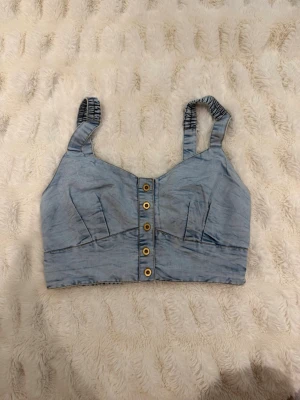 Ljusblå denim linnetopp med knappar - Trendig ljusblå linnetopp i denim-look med breda axelband och snygga knappar framtill. Toppen har en croppad passform och markerade sömmar för en cool och avslappnad stil. Perfekt att matcha med höga jeans eller kjol.