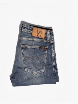 Blå smala jeans från Nudie Jeans - Säljer ett par klassiska blå jeans från Nudie Jeans med smal passform och fem fickor. Jeansen har snygga kontrastsömmar, läderpatch med logga bak och är gjorda i mjuk denim. Perfekta till en avslappnad stil.