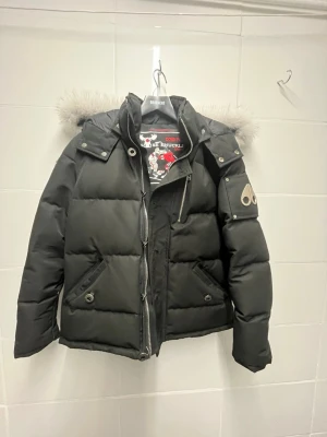 Svart pufferjacka med fuskpäls från Moose Knuckles - Snygg svart pufferjacka från Moose Knuckles med avtagbar huva och stor fuskpäls. Jackan har dragkedja, tryckknappar, två snedställda fickor och en logga på ärmen. Perfekt för kalla vinterdagar och riktigt trendig look.