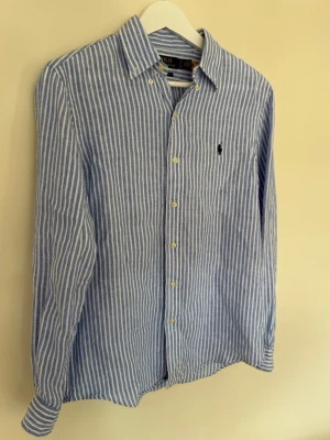 Blåvit randig skjorta från Polo Ralph Lauren - Klassisk blå och vit randig skjorta från Polo Ralph Lauren med lång ärm och knappar framtill. Skjortan har broderad logga på bröstet och är tillverkad i ett lätt och luftigt material, perfekt för en stilren look.
