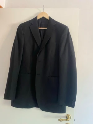 Svart kavaj från Dressmann D54 short  - Stilren svart kavaj från Dressmann med klassisk passform och två framfickor. Kavajen har smala slag och knäppning framtill. Perfekt för en dressad look. Materialet är slätt och fodret är svart.