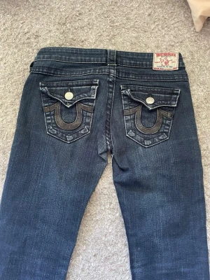 Mörkblå jeans True Religion - Säljer ett par lågmidjadd bootcut jeans från True Religion. Deras märke på fickorna är ”glitter”. Använda men inte slitna, omgjorda från straight till bootcut. Strl 26.