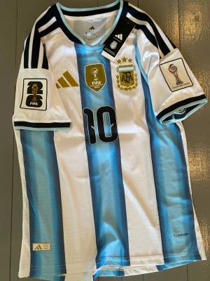 Argentina matchtröja Adidas nr 10 - Säljer en officiell Argentina fotbollströja från Adidas med nummer 10. Tröjan har klassiska blå och vita ränder, guldiga detaljer, FIFA World Champions märke och AFA-emblem. Korta ärmar med svarta och ljusblå detaljer. Perfekt för fotbollsfans!