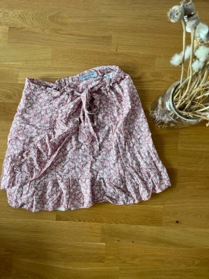 Blommig kortkjol från Montpoc - Söt kortkjol från Montpoc i ljusrosa med vitt blommönster. Kjolen har volangkant nertill och en knytning i midjan för extra detalj. Perfekt för varma dagar och ger en romantisk vibe till din outfit.