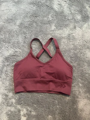 Vinröd sport-bh med korsade band - Vinröd sport-bh med ribbad resår och korsade axelband i ryggen. Bh:n har en sömlös design och bred kant nedtill för extra stöd. Perfekt för träning eller chill. Materialet känns mjukt och stretchigt.
