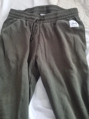 Olivgröna mjukisbyxor från Andy - Olivgröna mjukisbyxor från Andy med resår och dragsko i midjan. Byxorna har en avslappnad passform och är tillverkade i mjukt material, perfekta för chill dagar. Liten vit logolapp på höger sida upptill.