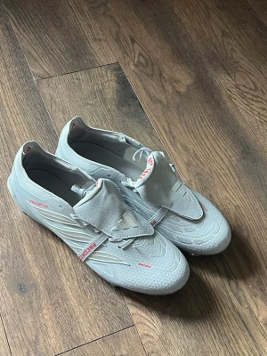 Adidas Predator vita fotbollsskor - Säljer ett par stilrena Adidas Predator fotbollsskor i vitt med röda detaljer och klassiska tre ränder på sidan. Skorna har snörning med täckt plös och är tillverkade i syntetmaterial för bästa grepp och känsla på planen. Perfekta för dig som vill sticka ut på matchen.