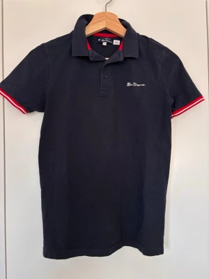 Mörkblå pikétröja från Ben Sherman, 12-13 år - Klassisk mörkblå pikétröja från Ben Sherman med vit logga på bröstet och röda detaljer på krage och ärmslut. 
