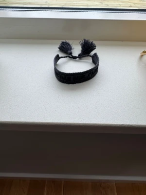 Svart tygarmband med tofsar - Stilrent svart armband i tyg med bred design och två dekorativa tofsar som knyts. Perfekt accessoar för att ge din outfit en cool och avslappnad touch. Jag fick det så vet ej något om vart det är ifrån eller vart de är köpt.