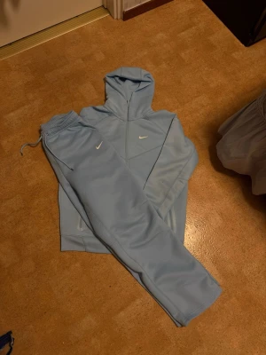 Ljusblå Nike loungewear-set - Snyggt ljusblått loungewear-set från Nike med matchande hoodie och byxor. Setet har diskreta vita Nike-loggor och texten A.W.R. på byxbenet. Mjukt material och avslappnad passform, perfekt för chill dagar hemma.