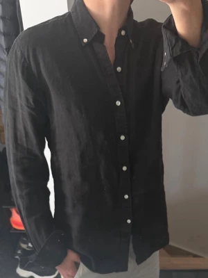 Svart linneskjorta med knappar - Stilren svart skjorta i linne från John Henric med klassisk krage och vita knappar hela vägen. Skjortan har lång ärm och en avslappnad passform, perfekt för en clean och enkel look. Storlek 39 vilket motsvarar m