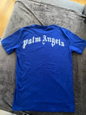 Blå Palm Angels t-shirt - Snygg blå t-shirt från Palm Angels med vit logga i gotisk stil på ryggen och mindre tryck vid halsen fram. Klassisk passform med rund hals och korta ärmar. Perfekt för dig som gillar streetwear och vill sticka ut. Inga skador eller hål 