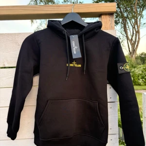 Svart hoodie från Stone Island - Tja! Säljer en Snygg svart hoodie från Stone Island med gul logga på bröstet och stort tryck på ryggen i vitt, gult och grått, eftersom den var för liten för mig, därmed är den inte använd! 