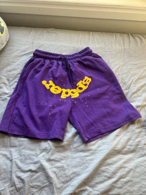 Lila shorts med gul Sp5der-logga - Säljer ett par lila shorts från Sp5der med stor gul logga framtill. Shortsen har resårmidja med snörning och en bakficka. Tillverkade i mjuk bomull, perfekta för sommardagar. Snygg streetwear vibe och loose fit.