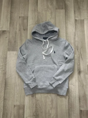 Grå hoodie från Polo Ralph Lauren - Snygg grå hoodie från Polo Ralph Lauren med klassisk huva och vita dragsnören. Tröjan har en känguruficka framtill och den ikoniska lilla loggan broderad på bröstet. Tillverkad i mjukt bomullsmaterial och har ribbade muddar vid ärmar och nederkant.