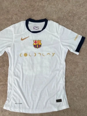 Vit FC Barcelona matchtröja Nike - Säljer en vit FC Barcelona matchtröja från Nike med klubbmärke och guldfärgade detaljer. Tröjan har mörkblå krage och ärmslut, tryck med Lamine Yamal och nummer 10 på ryggen. Materialet är lätt och andas, perfekt för fotbollsträning.