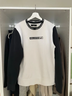Vit och svart Hollister sweatshirt - Snygg sweatshirt från Hollister i vitt med svarta ärmar och svart ribbad krage med Hollister-tryck. Klassisk logga på bröstet och mjukt material, perfekt för en avslappnad streetwear-look. Tröjan har en rak passform och långa ärmar.
