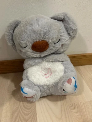Grå koala mjukisdjur med sång, andning, och belysning - Supergulligt mjukisdjur i form av en koala med grå päls, vitt mageparti och brunt nosparti. Koalan har broderade ögon och små rosa detaljer på kinderna. Tassarna har blå och rosa detaljer. Perfekt som kramkompis eller dekoration.