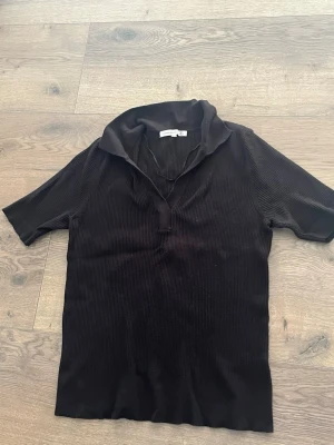 Svart ribbad topp med krage - Svart ribbad topp med korta ärmar och bred krage. Toppen har en djupare v-ringning och är tillverkad i ett mjukt, stretchigt material. Perfekt för en stilren och avslappnad look.