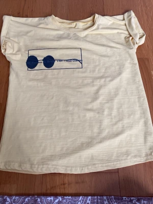 Gul t-shirt med tryck och rund hals - Ljusgul t-shirt i bomull med korta ärmar och ett tryck av blå solglasögon och text på bröstet. T-shirten har en avslappnad passform och rund halsringning. Perfekt för en chill och enkel stil.