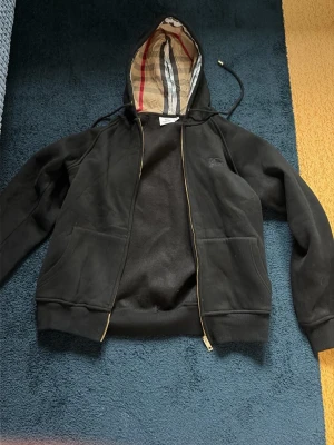 Svart hoodie från Burberry - Snygg svart hoodie från Burberry med dragkedja och klassiskt rutigt foder i huvan. Hoodien har två stora fickor framtill och justerbar dragsko i huvan. Perfekt för en avslappnad och trendig look.