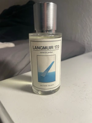 Langmuir 17.0 Extrait de Parfum - En stilren parfymflaska från One Bold Chemist med doften Langmuir 17.0. Flaskan är cylindrisk i genomskinligt glas med en silverfärgad metallkork. Etiketten är vit med svart text och en blå grafisk illustration. 