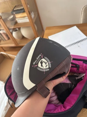 Svart ridhjälm från Fouganza - Snygg svart ridhjälm från Fouganza med vit detalj och Horse Riding-märke på sidan. Hjälmen har ventilerande hål upptill och justerbart hakband. Perfekt för dig som gillar hästar och vill ha en stilren look i stallet.