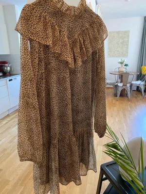 Leopardmönstrad volangklänning - Säljer en luftig leopardmönstrad klänning med volangdetaljer och hög krage. Klänningen har lång ärm och är tillverkad i ett tunt, transparent material som ger en snygg och trendig look. Perfekt för dig som vill sticka ut med ett djurmönster.