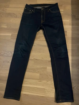 Mörkblåa jeans från Nudie - Snygga mörkblåa jeans från Nudie med klassiska kontrastsömmar och femficksdesign. Jeansen har en smal passform och är tillverkade i stretchigt denimtyg för extra komfort. Perfekta för en trendig och avslappnad look.