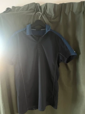 Golfpike Galvin Green - Sportig mörkblå pikétröja från Galvin Green med blå detaljer på axlar och ärmar. Tröjan har klassisk krage, knäppning med tre knappar och vit söm längs sidorna. Tillverkad i ett ventilerande funktionsmaterial, perfekt för golf eller annan träning.