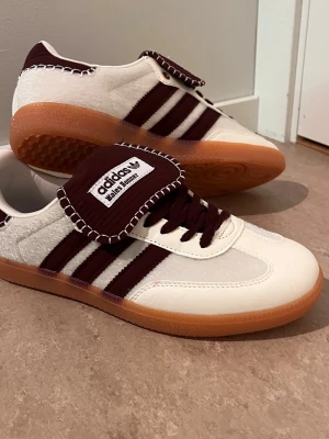 Adidas Samba OG vit vinröd sneakers - Adidas Samba OG sneakers i vitt och vinrött med klassiska tre ränder på sidan, brun gummisula och bred plös med Adidas-logga. Ovandelen är i mocka och läder, detaljer i vinrött och vita sömmar. Perfekt för dig som gillar retrostil och streetwear.