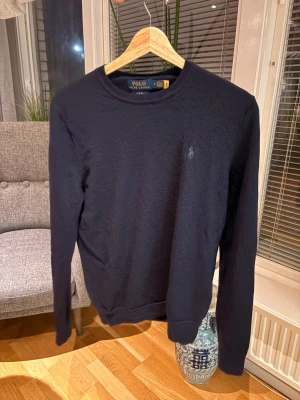 Mörkblå merinoullströja Polo Ralph Lauren - Slim fit mörkblå tröja från Polo Ralph Lauren i 100% merinoull. Klassisk rund halsringning, diskret broderad logga på bröstet och långa ärmar. Perfekt för dig som gillar stilren och tidlös design med en lyxig känsla. 