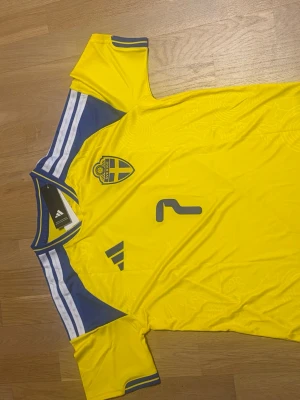 Sveriges vm tröja bergvall M - Säljer en gul svensk landslagströja från Adidas med blå detaljer och vita ränder på axlarna. Tröjan har nummer 2 och namnet 'Bergvall' på ryggen samt Sveriges landslagsemblem på bröstet. Perfekt för dig som vill stötta Sverige på match!