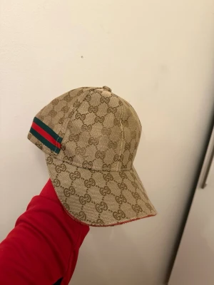 Beige keps från med GG-mönster - Säljer en beige keps från  med klassiskt GG-monogram över hela kepsen. På sidan finns en grön och röd rand, samt justerbart spänne bak med G-detalj i metall. Kepsen är i canvasmaterial och har böjd skärm.
