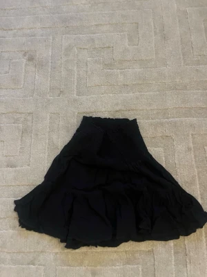 Svart volangkjol med resårmidja - Snygg svart kjol med volanger och bred resår i midjan. Kjolen har en luftig och flowy passform, perfekt för en trendig och avslappnad stil. Materialet känns lätt och bekvämt mot huden.