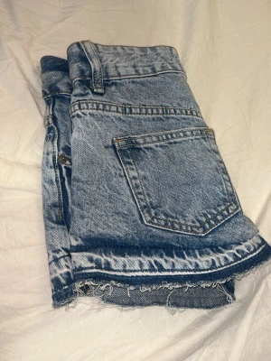 Lågmidjde Blå jeansshorts med rå kant - Säljer ett par klassiska blå jeansshorts i denim med låg midja och rå, fransig kant nedtill. Shortsen har bakficka och tydliga sömmar för en snygg vintagekänsla. Perfekta till sommaren och lätta att matcha med olika stilar.