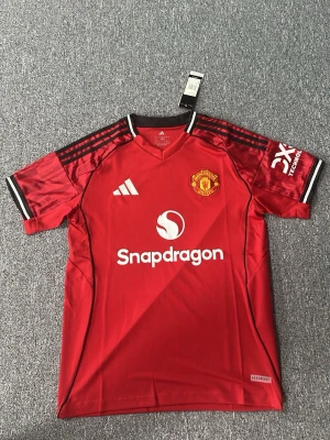 Manchester United matchtröja röd - Snygg röd Manchester United matchtröja från Adidas med klubbmärke på bröstet och Snapdragon som sponsortryck. Tröjan har svarta och vita detaljer på ärmarna, klassiska Adidas-ränder på axlarna och är tillverkad i ett lätt, ventilerande material.