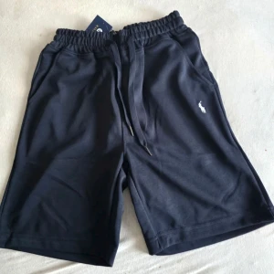 Marinblå shorts från Polo Ralph Lauren - Snygga marinblå shorts från Polo Ralph Lauren med elastisk midja och snörning. Klassisk logga broderad i vitt på ena benet. Tillverkade i mjuk bomull, perfekta för avslappnade dagar. Två sidofickor och en bekväm passform.