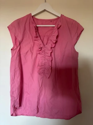 Rosa somrig  blus med volangdetalj - Snygg klarrosa blus i luftig bomull med korta ärmar och volangdetalj framtill. Perfekt nu i sommar!💞 har för mig den är från H&M