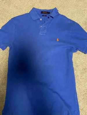 Blå pikétröja från Ralph Lauren - Klassisk blå pikétröja från Ralph Lauren med orange broderad logga på bröstet. Slim fit-modell med korta ärmar och krage. Tillverkad i bomull för en skön och luftig känsla. Perfekt för en avslappnad men ändå stilren look.