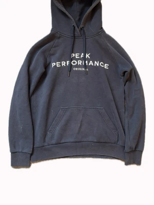 Peak Performance mörkblå hoodie  - Mörkblå hoodie från Peak Performance med vit text på bröstet. Klassisk känguruficka framtill, ribbade muddar och justerbar huva med snören. Storlek S 