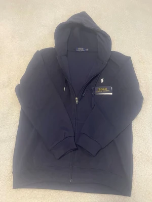 Mörkblå hoodie från Polo Ralph Lauren - Snygg mörkblå hoodie från Polo Ralph Lauren med dragkedja framtill och klassisk vit logga broderad på bröstet. Hoodien har justerbar huva med snören och ribbade muddar vid ärmar och nederkant. Tillverkad i mjukt bomullsmaterial för en skön känsla.