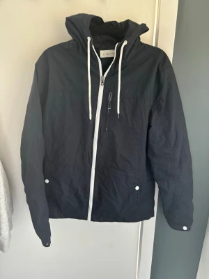 Svart vindjacka från pier one - Snygg svart vindjacka från pier one med vit dragkedja och vita snören i huvan. Jackan har en bröstficka med dragkedja och två sidofickor med knappar. Perfekt för blåsiga dagar och enkel att matcha med olika outfits.