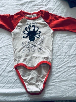 Body från Mini Rodini med bläckfisk - Säljer en söt body från Mini Rodini med röda ärmar och kantband. Framsidan har ett tryck med en svart bläckfisk, vågor och texten 'A LA MER!'. Tillverkad i mjuk bomull, perfekt för små barn.