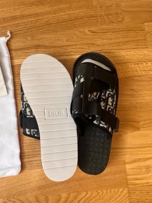 Svarta  sandaler med monogram - Säljer ett par svarta sandaler från med klassiskt monogrammönster i vitt och svart. Sandalerna har två breda remmar med justerbara spännen och vit räfflad sula. Perfekta för dig som vill ha en lyxig och trendig look.