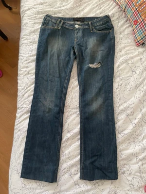 Blå bootcut jeans  - Snygga blå bootcut jeans med slitningar och ett hål på låret för en trendig look. Klassisk femficksmodell med låg midja och detaljerade bakfickor. Perfekta för dig som gillar en avslappnad men ändå stilsäker stil. Jag har sytt upp den så den är lite kortare än vad som står. Finns ingen storlek men skulle säga M/L
