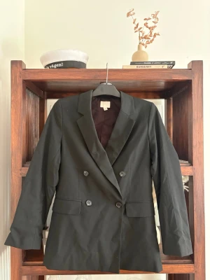 Svart dubbelknäppt kavaj - Stilren svart kavaj med dubbelknäppning och klassisk krage. Långa ärmar, två fickor framtill och slät finish. Perfekt för en trendig och uppklädd look. Fodrad insida för extra komfort.