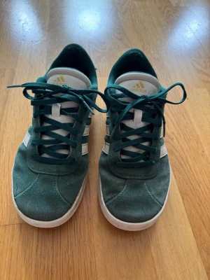 Gröna Adidas sneakers i mocka - Snygga gröna Adidas sneakers i mocka med klassiska vita ränder på sidorna och vit sula. Skorna har lågt skaft och gröna skosnören. Perfekta för dig som gillar retrostil och vill ha något bekvämt till vardags. Adidas-logga på plösen och hälen. Rengjorda!