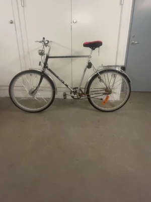 Klassisk herrcykel med röd sadel - Säljer en klassisk herrcykel i svart och silver med röd sadelöverdrag. Cykeln har raka styren, fram- och baklykta samt pakethållare bak. Utrustad med reflexer och kedjeskydd. Perfekt för dig som vill ha en enkel och funktionell cykel.